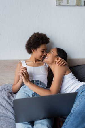 cheerful african american lesbian couple watching movie on laptop and holding hands in bedroom