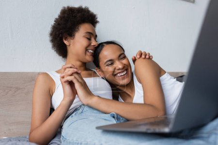 joyful african american lesbian couple watching movie on laptop while holding hands