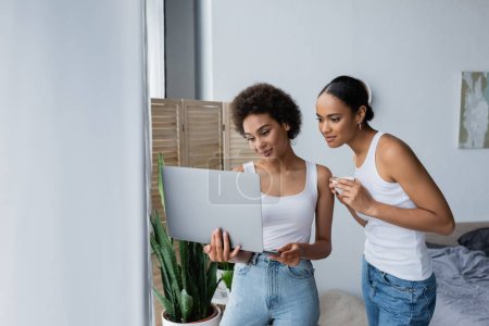 Téléchargez les photos : Africaine américaine lesbienne femme à l'aide d'ordinateur portable tandis que petite amie tenant tasse de café - en image libre de droit
