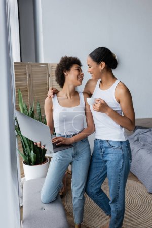 cheerful african american lesbian woman using laptop while girlfriend holding cup of coffee 