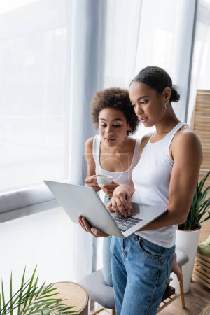 african american lesbian woman using laptop while curly girlfriend holding cup of coffee 