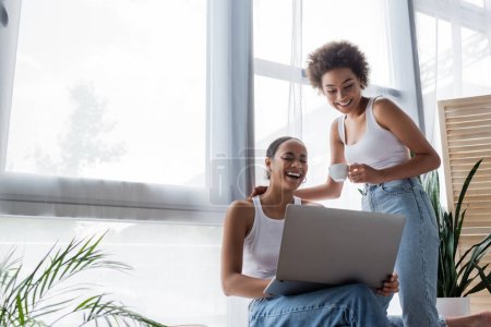 junge afrikanisch-amerikanische lesbische Frau mit Laptop in der Nähe freudige Freundin hält Tasse Kaffee 