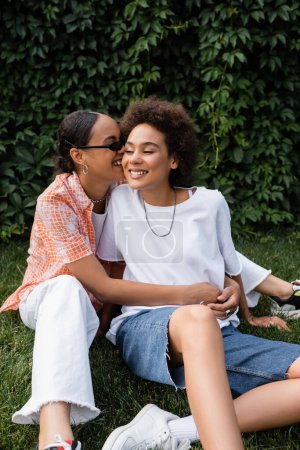 elegante africano americano lesbiana mujer en gafas de sol abrazando feliz novia mientras sentado en el césped 