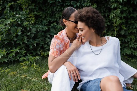 fröhliche afrikanisch-amerikanische lesbische Frau mit Sonnenbrille umarmt glückliche Freundin, während sie auf dem Rasen sitzt 