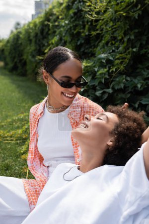 freudige afrikanisch-amerikanische lesbische Frau mit Sonnenbrille, die auf dem Rasen sitzend eine glückliche Freundin ansieht 