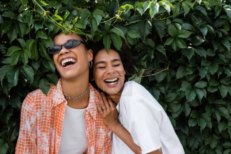 happy african american lesbian women laughing near green bush in park 