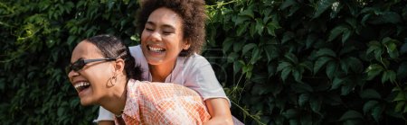 Photo pour Heureux afro-américain lesbienne femme embrasser excité copine dans lunettes de soleil rire dans vert parc, bannière - image libre de droit