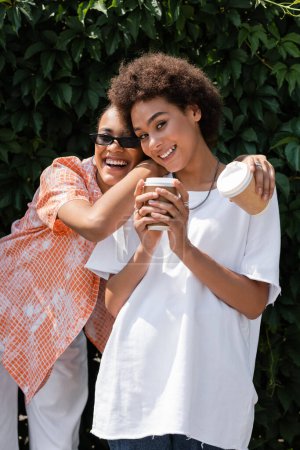 sonriente afroamericana lesbiana mujer en gafas de sol abrazando rizado novia y sosteniendo café para ir