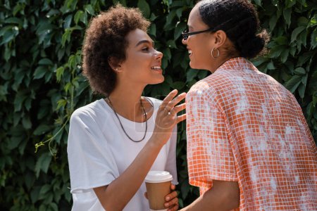 happy african american lesbian woman in sunglasses looking at curly girlfriend with  coffee to go