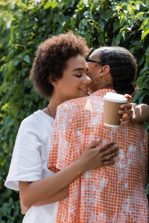 happy african american lesbian woman hugging girlfriend and holding coffee to go