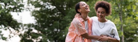 joyful african american lesbian woman hugging happy girlfriend and laughing in green park, banner