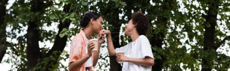 joyful african american lesbian woman holding coffee to go and kissing hand of girlfriend in park, banner