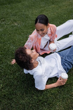 Téléchargez les photos : Vue aérienne de joyeuses femmes lesbiennes afro-américaines tenant des tasses en papier et assises sur l'herbe dans le parc - en image libre de droit