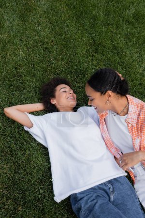 Ansicht der glücklichen afrikanisch-amerikanischen lesbischen Frau, die ihr Smartphone in der Nähe ihrer Freundin hält, die im Gras liegt 