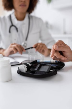 abgeschnittene Ansicht eines Mannes mittleren Alters mit Diabetes, der auf ein Glukometergerät in der Nähe eines amerikanischen Arztes zeigt