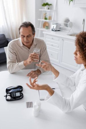 curly african american doctor gesturing while talking with middle aged patient holding glass of water 