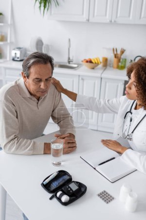 Photo for Young african american doctor in white coat calming sad middle aged man with diabetes - Royalty Free Image