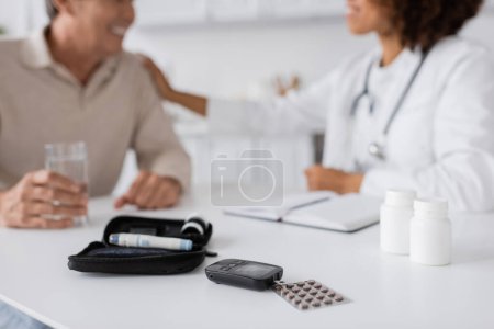 Diabetes-Set mit Glukosemessgerät und Lanzettstift-Geräten in der Nähe von afrikanisch-amerikanischem Arzt und Patient auf verschwommenem Hintergrund 