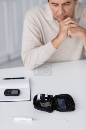 Mann mittleren Alters betrachtet Diabetes-Set mit Glukosemessgerät und Lanzettenstift auf dem Tisch 