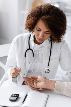 médecin afro-américain bouclé prélèvement d'échantillon de sang de patient mature avec stylo lancette près du mètre de glucose sur la table 