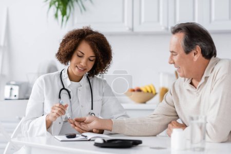 Photo for Smiling african american doctor taking blood sample of mature patient with lancet pen - Royalty Free Image