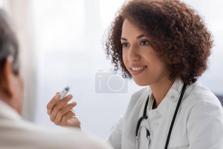 smiling african american doctor holding lancet pen device near blurred man 
