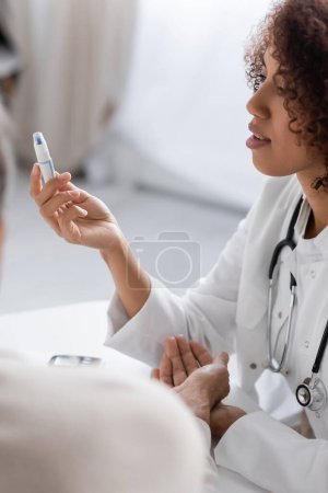 young and curly african american doctor holding lancet pen device near blurred patient 