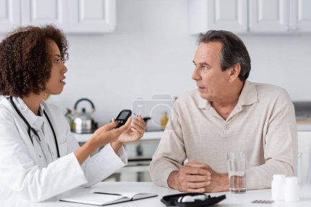 curly african american doctor holding glucometer device near middle aged patient
