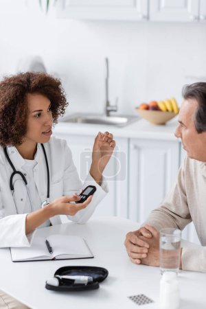 médico afroamericano rizado sosteniendo dispositivo de glucosímetro y hablando con paciente de mediana edad