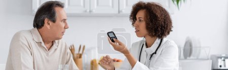 Téléchargez les photos : Vue latérale du médecin afro-américain bouclé tenant le dispositif de glucomètre et parlant avec un patient d'âge moyen, bannière - en image libre de droit