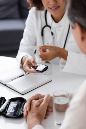 cropped view of african american doctor in white coat holding glucometer device and test strip near middle aged patient