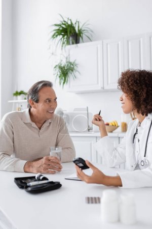curly african american doctor holding glucometer near smiling middle aged patient 