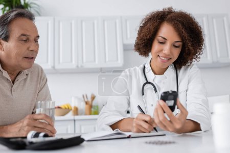 lächelnder afrikanisch-amerikanischer Arzt mit Glukometer und Notizbuch in der Nähe eines Patienten mittleren Alters 