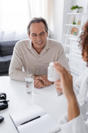 smiling middle aged man with diabetes looking at bottle with pills in hand of african american doctor 