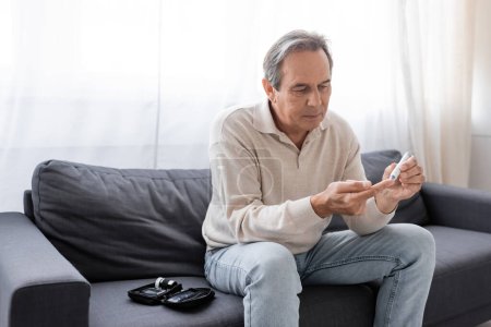 Mann mittleren Alters nimmt Blutprobe mit Lanzettstift, während er auf Couch im Wohnzimmer sitzt 