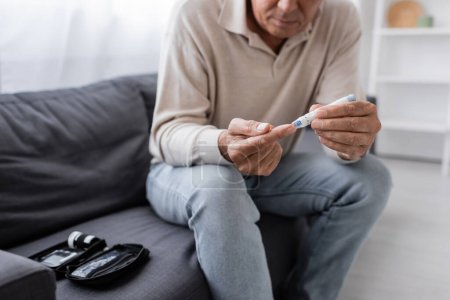 vista recortada del hombre de mediana edad con diabetes tomando muestras de sangre mientras está sentado en el sofá en la sala de estar 