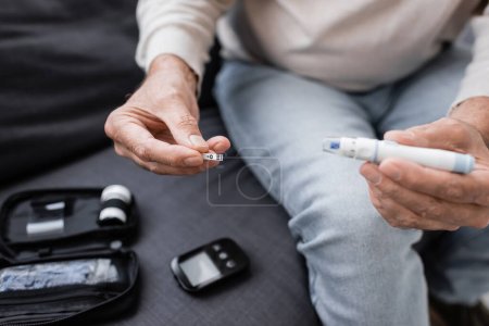 Photo for Cropped view of middle aged man with diabetes holding lancet pen and test strip while sitting on couch in living room - Royalty Free Image