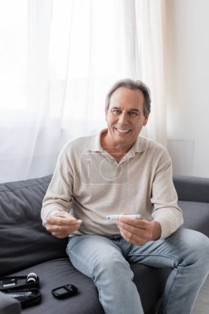 Photo for Cheerful mature man with diabetes holding lancet pen and test strip while sitting on couch in living room - Royalty Free Image