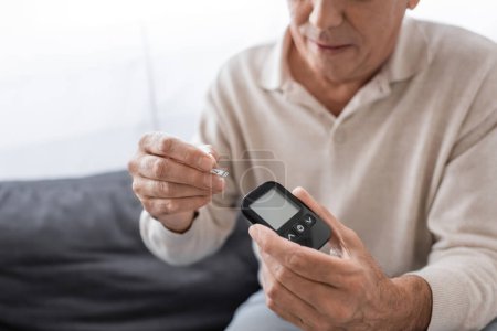 Téléchargez les photos : Vue recadrée de l'homme d'âge moyen avec diabète tenant appareil de mesure de glucose et bande d'essai - en image libre de droit