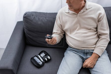 Teilansicht eines Mannes mittleren Alters mit Diabetes, der ein Glukosemessgerät in der Hand hält und auf dem Sofa sitzt 