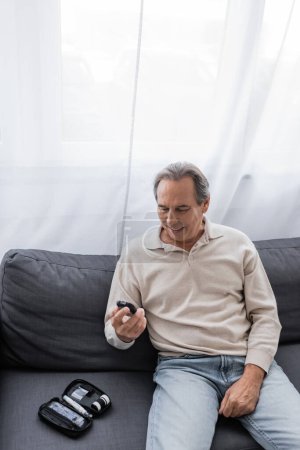 alegre hombre de mediana edad con diabetes sosteniendo el dispositivo medidor de glucosa y sentado en el sofá 