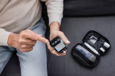 partial view of middle aged man with diabetes pointing at glucose meter device and sitting on sofa 
