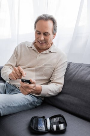 Téléchargez les photos : Homme d'âge moyen joyeux avec diabète pointant vers l'appareil de mesure de glucose et assis sur le canapé - en image libre de droit