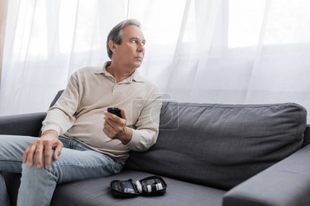 Téléchargez les photos : Homme d'âge moyen avec diabète tenant appareil de mesure de glucose et assis sur le canapé dans le salon - en image libre de droit