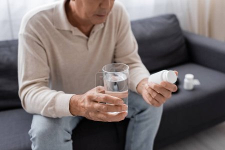 Teilansicht eines Mannes mittleren Alters mit Diabetes, der auf dem Sofa sitzt und ein Glas Wasser und Tabletten in der Hand hält 