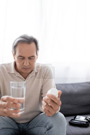 trauriger Mann mittleren Alters mit Diabetes, der ein Glas Wasser und Tabletten hält, während er auf dem Sofa sitzt 