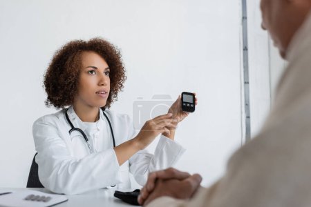 Téléchargez les photos : Médecin afro-américain bouclé tenant dispositif de glycomètre près de l'homme d'âge moyen avec diabète en clinique - en image libre de droit