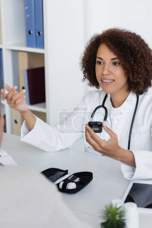 Téléchargez les photos : Médecin afro-américain joyeux tenant glucomètre et parlant avec un patient d'âge moyen atteint de diabète - en image libre de droit