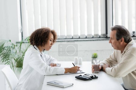 african american doctor writing on clipboard near mature patient with diabetes holding glass of water in clinic 