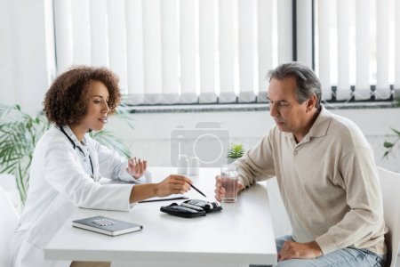 médecin afro-américain pointant vers le dispositif glucomètre avec stylo près d'un patient mature diabétique tenant un verre d'eau